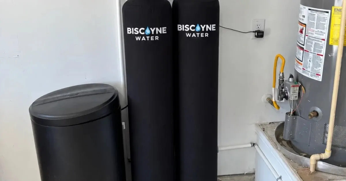 Water softener media tank and brine tank installed in a South Florida garage beside a water heater with visible limescale